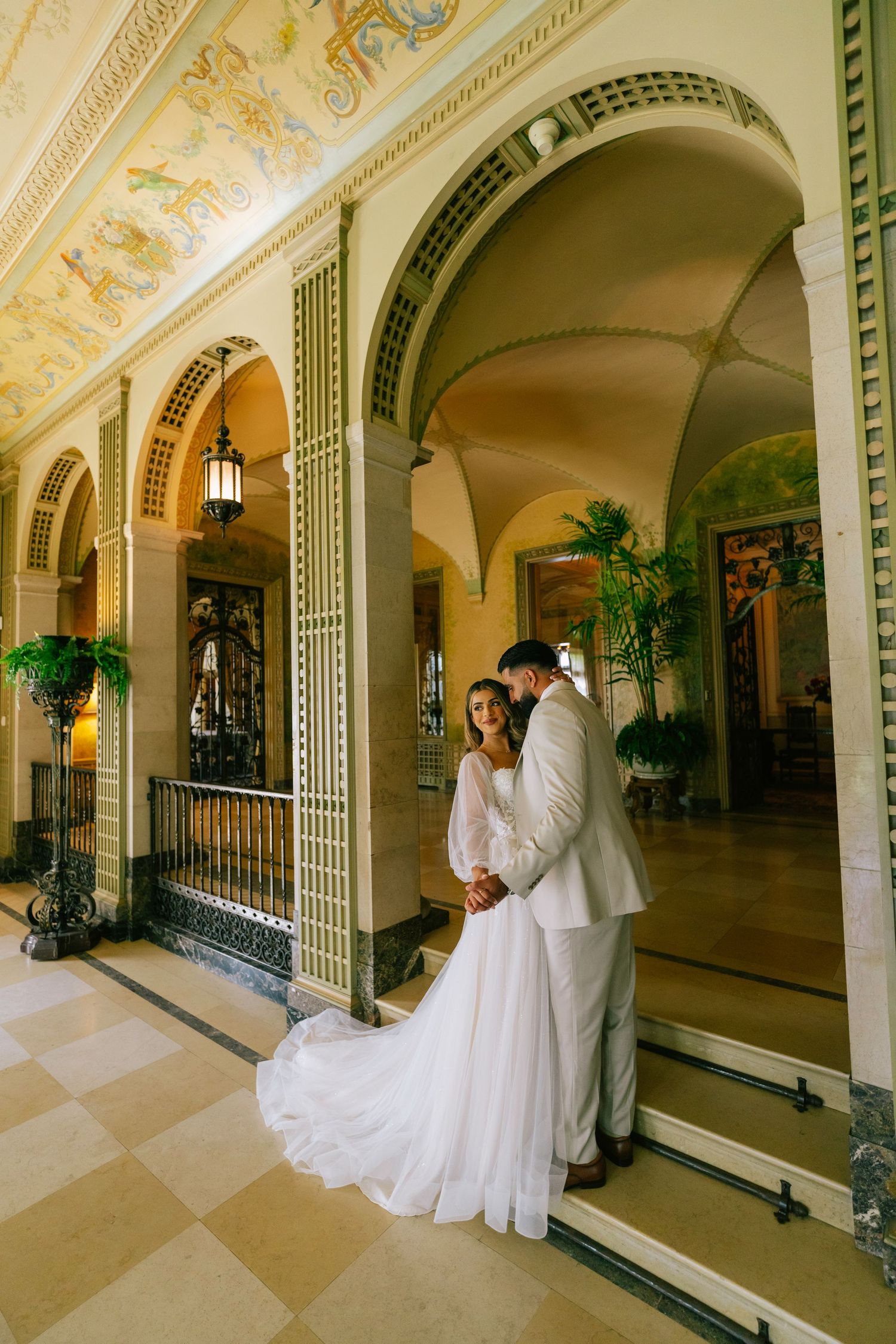 Elegant couple in white wedding attire embracing in an ornate historic hallway with arched columns and checkerboard floors.