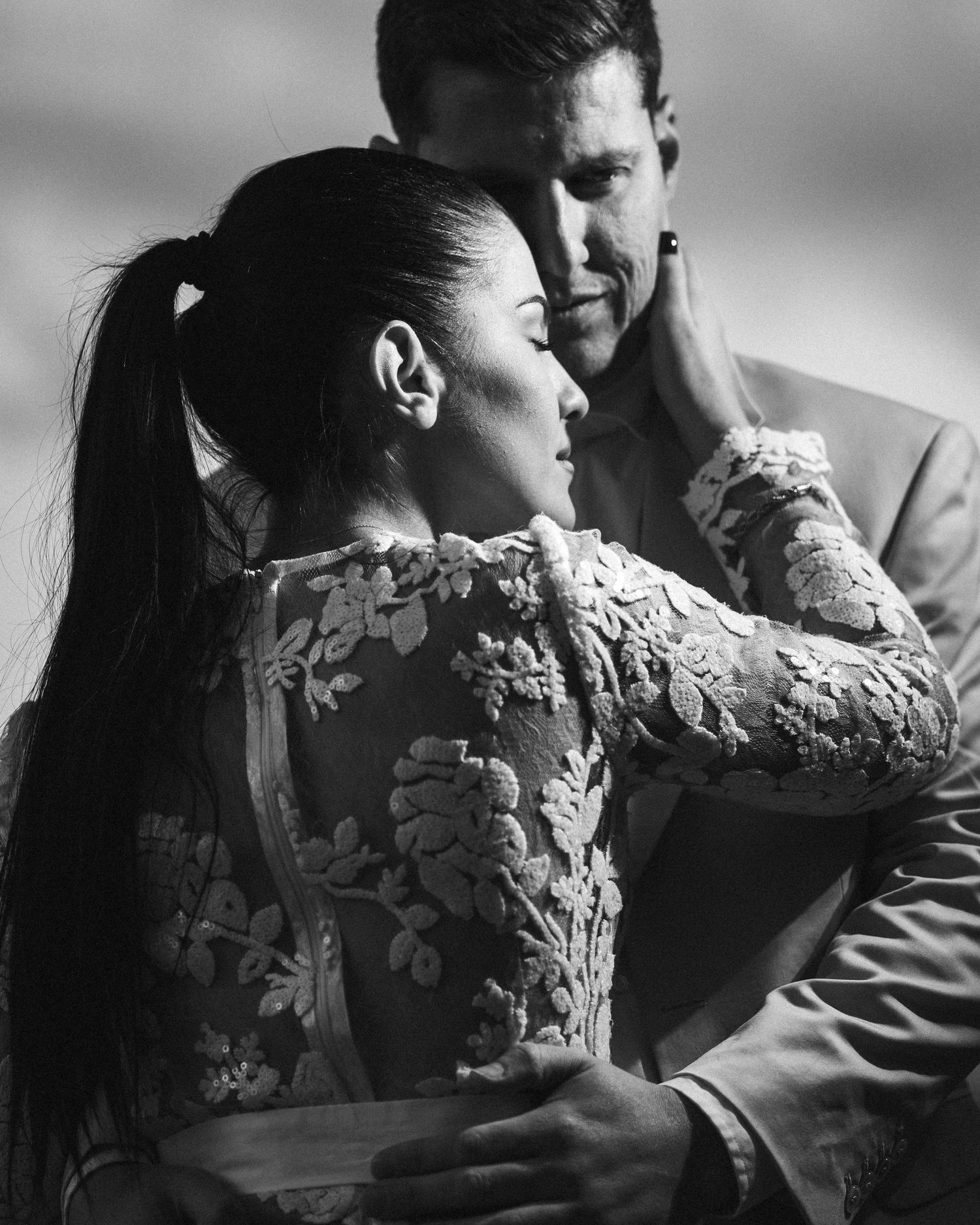 Black and white portrait of a couple in an intimate embrace with the woman wearing an ornate lace dress.