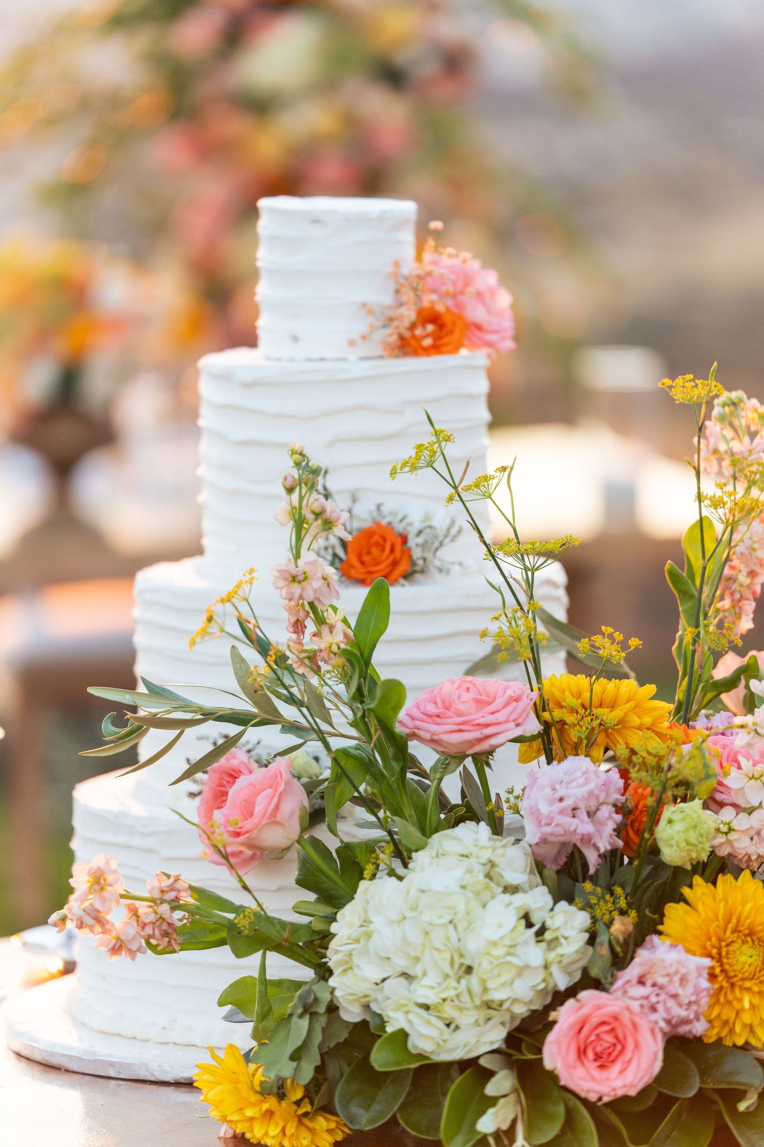 Elegant white tiered wedding cake decorated with pink, orange, and yellow fresh flowers. γαμήλια τούρτα.