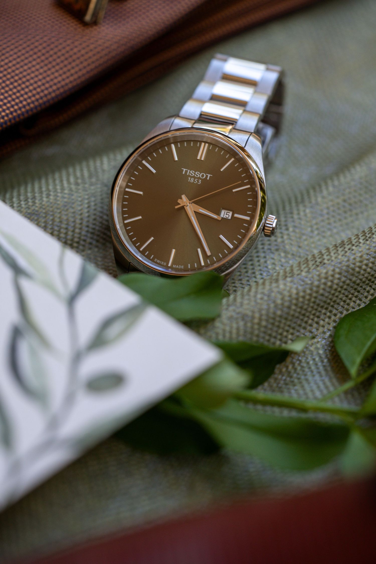 Silver Tissot watch with brown dial displayed on green fabric with leaves.