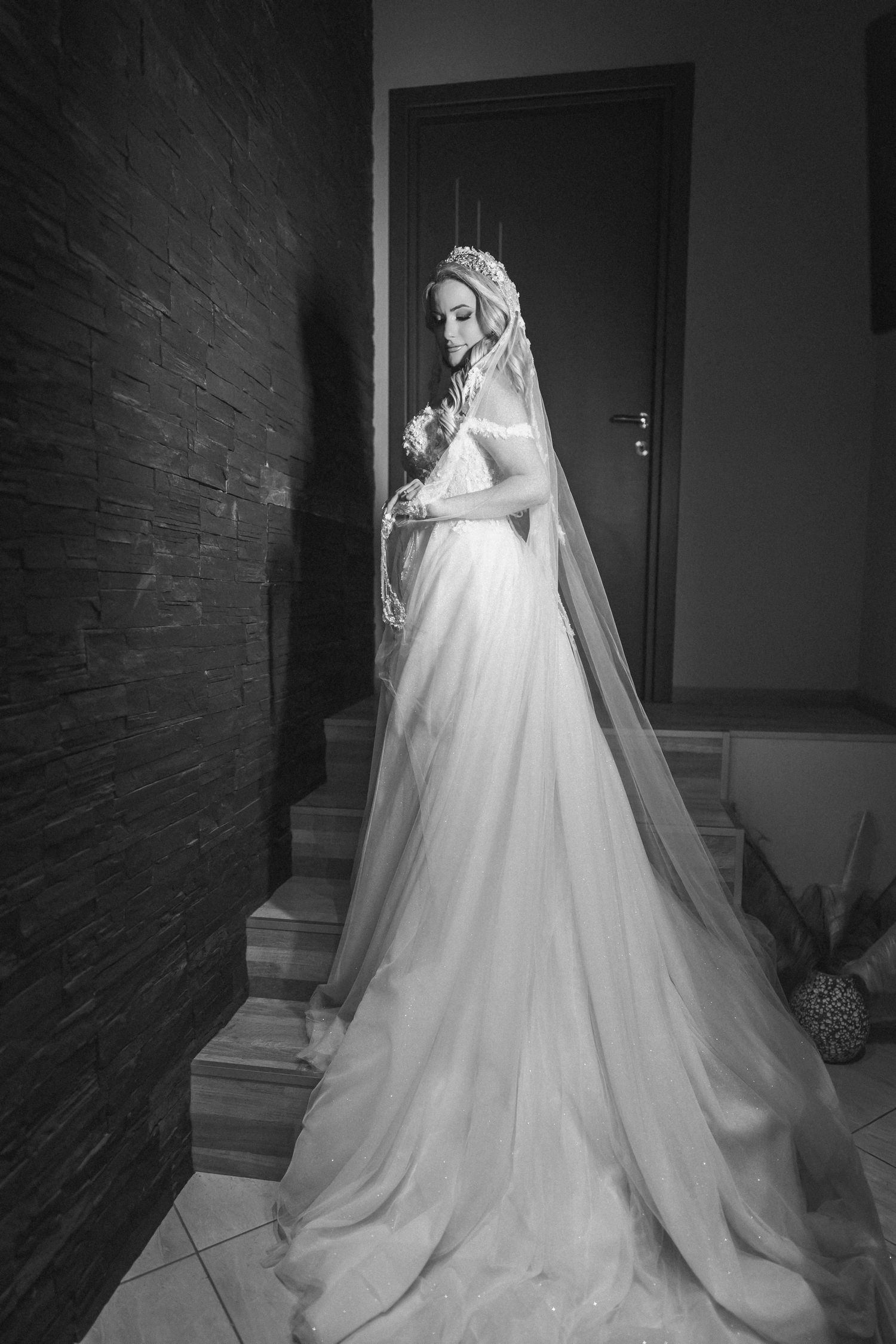 A bride in an elegant white wedding gown with long flowing train and veil poses on stairs against a brick wall.