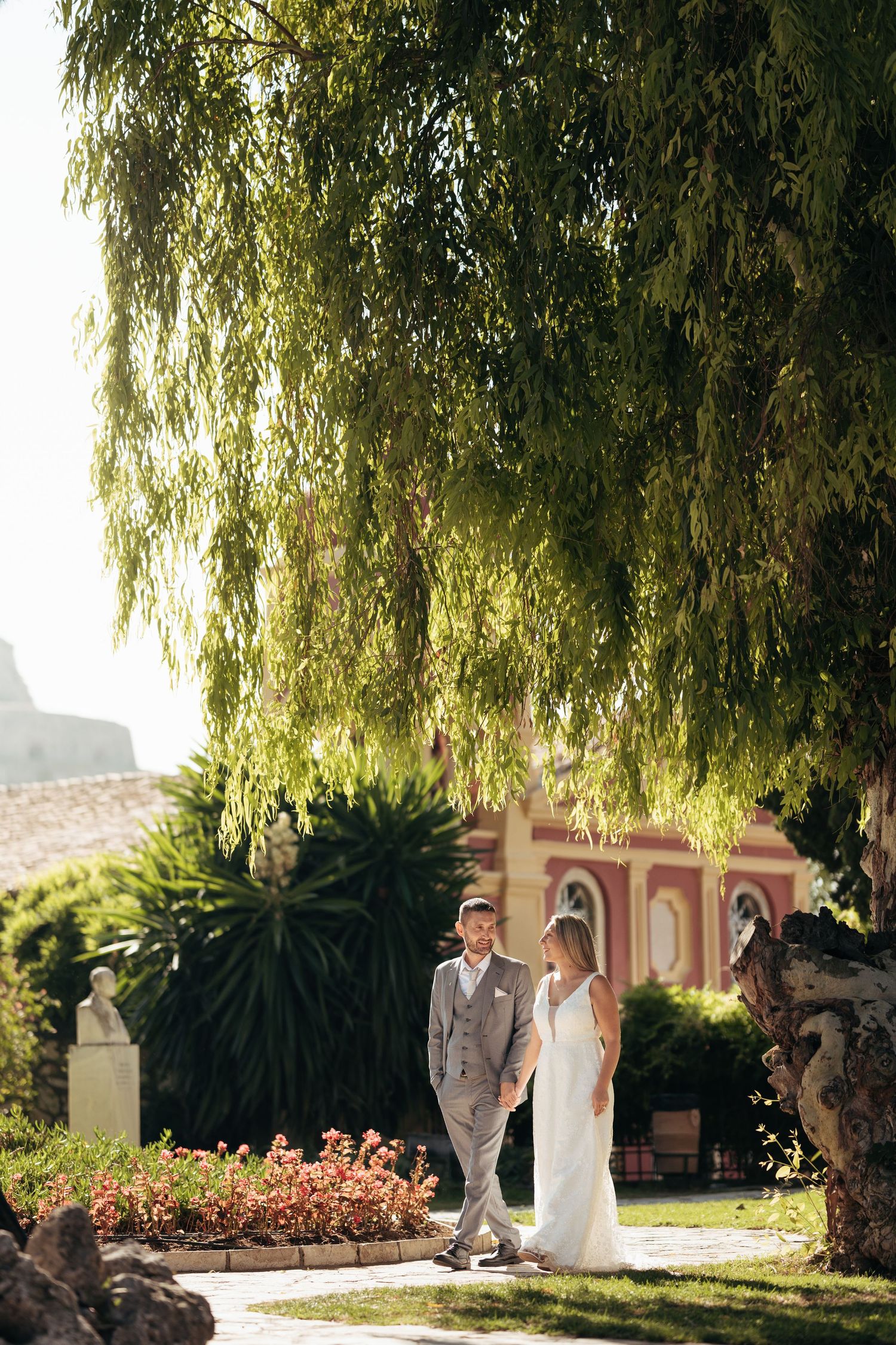 Wedding couple poses under drooping willow trees on a sunny garden estate.
