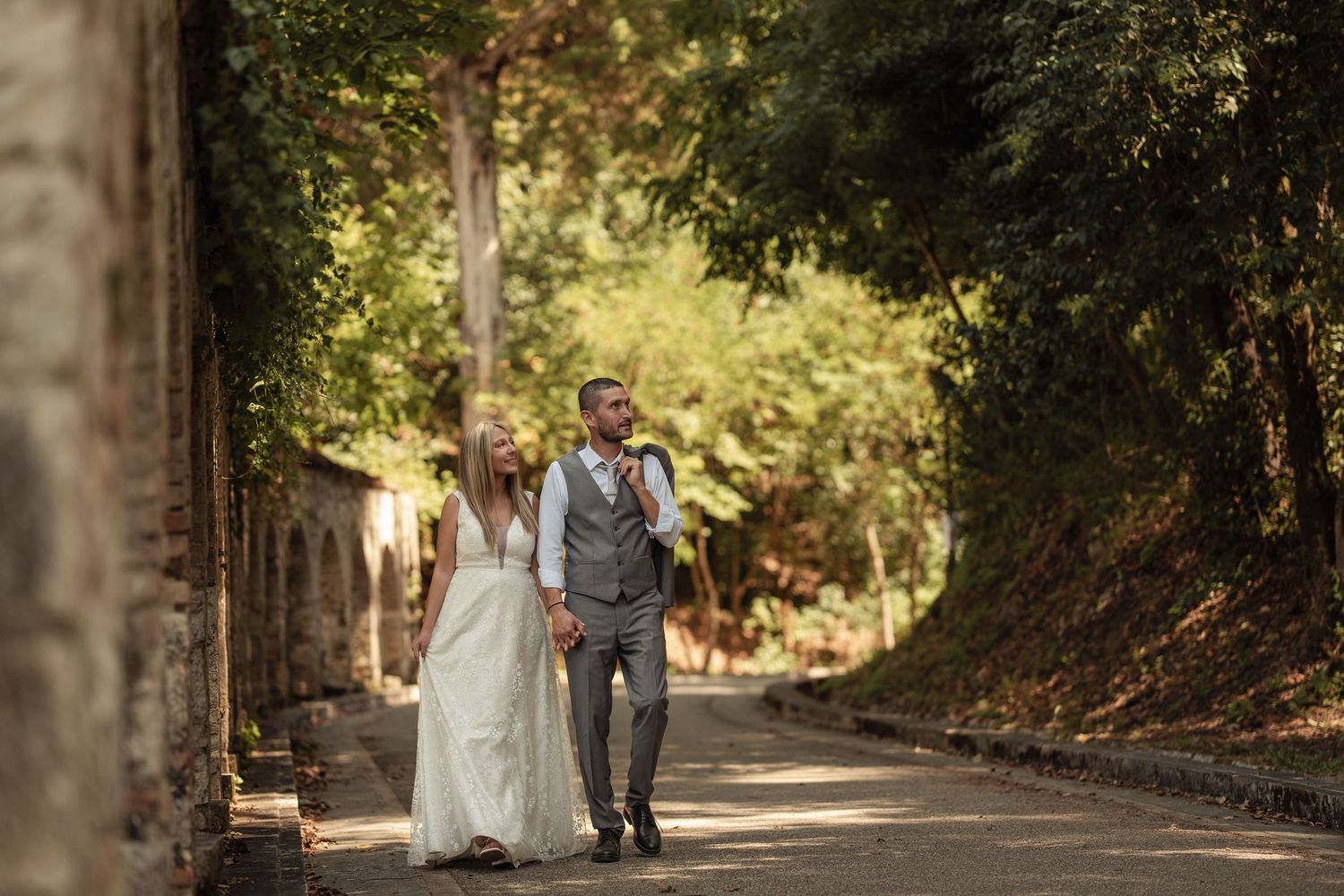 A couple walks hand in hand down a tree-lined path on their wedding day in warm sunlight.