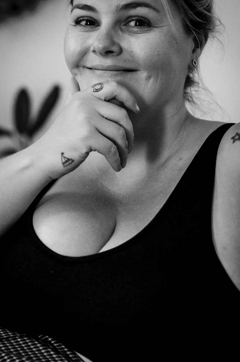 Black and white portrait of a person in a tank top smiling while resting their hand on their chin with a small tattoo visible.