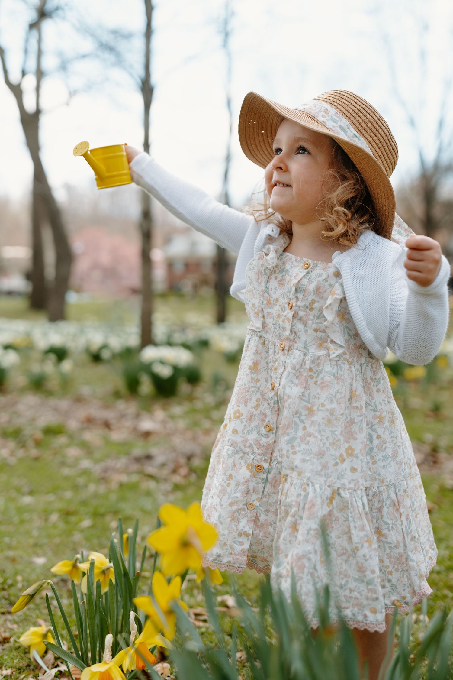 Child in floral dress and straw hat exploring spring daffodils in a garden setting.
