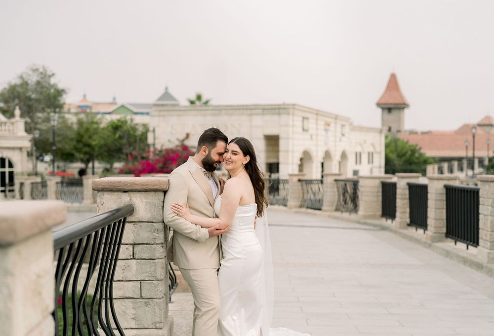 Romantic couple embraces on a stone bridge with elegant white buildings and a turret in the background.