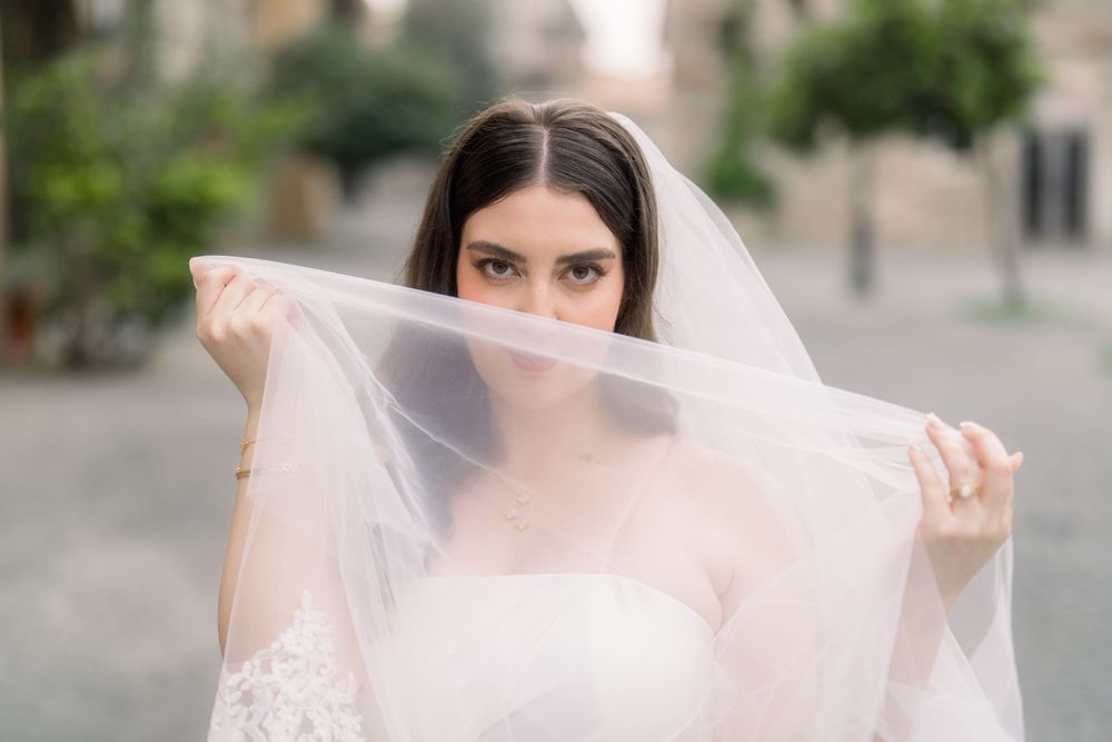 Ethereal image of a bride peeking through her delicate lace veil in a soft outdoor setting.