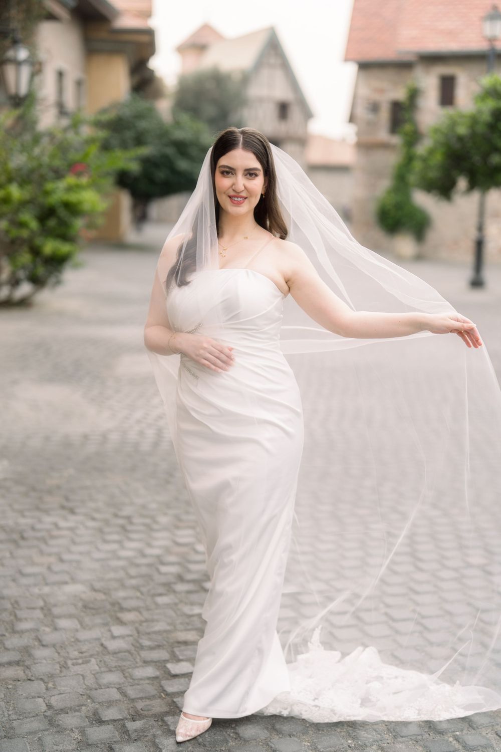 Elegant wedding dress with flowing veil and fitted silhouette photographed on cobblestone street with houses in background.