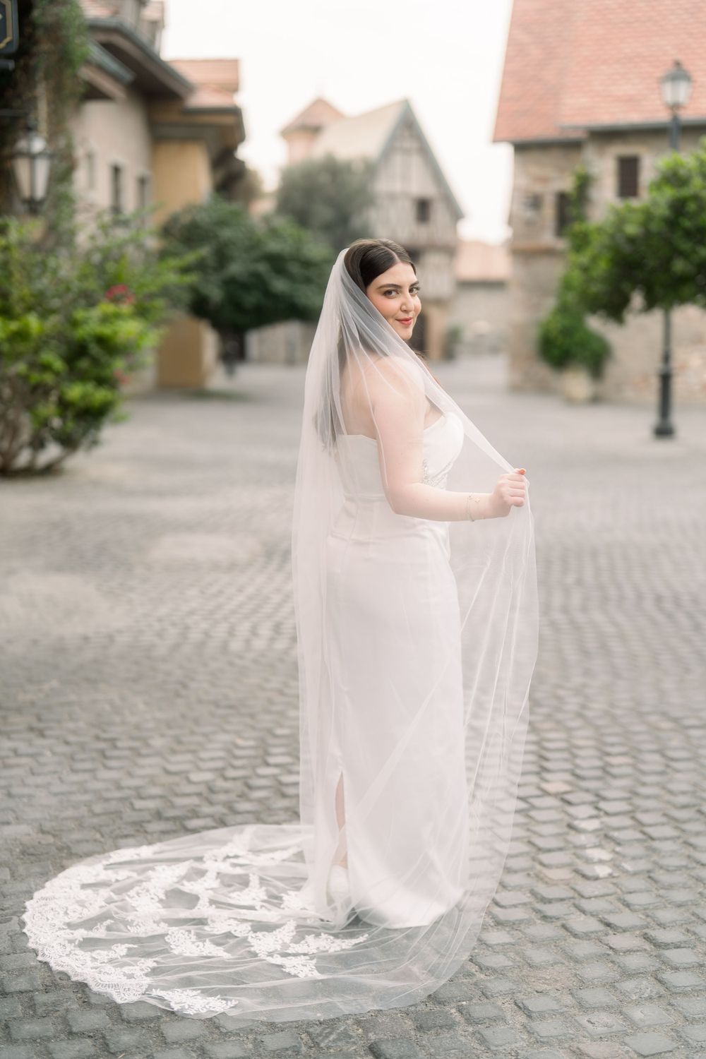 A bride in a sleek white gown and long lace-trimmed veil poses on a cobblestone street near traditional buildings.