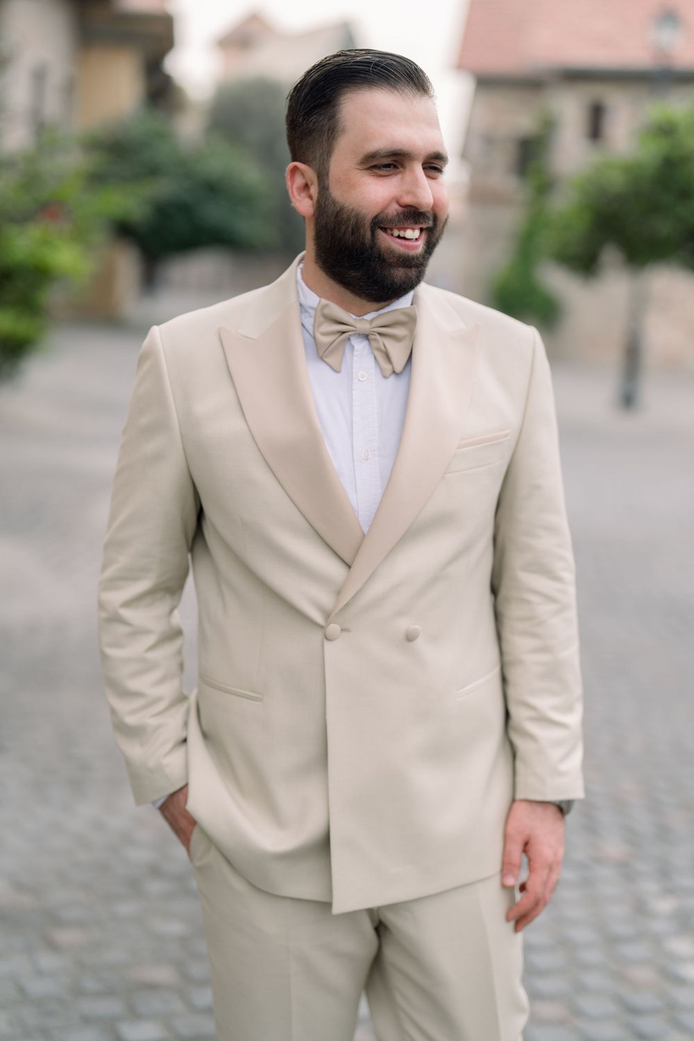 A person wearing a beige double-breasted suit with a bow tie smiles while standing outdoors on a cobblestone street.