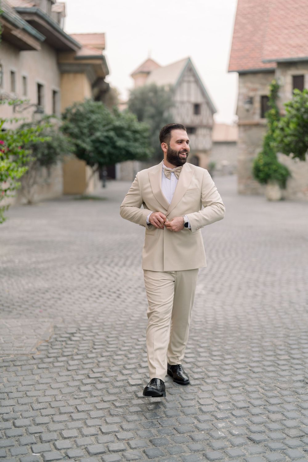 A person wearing a beige suit and bow tie stands on a cobblestone street in a historic European town setting.