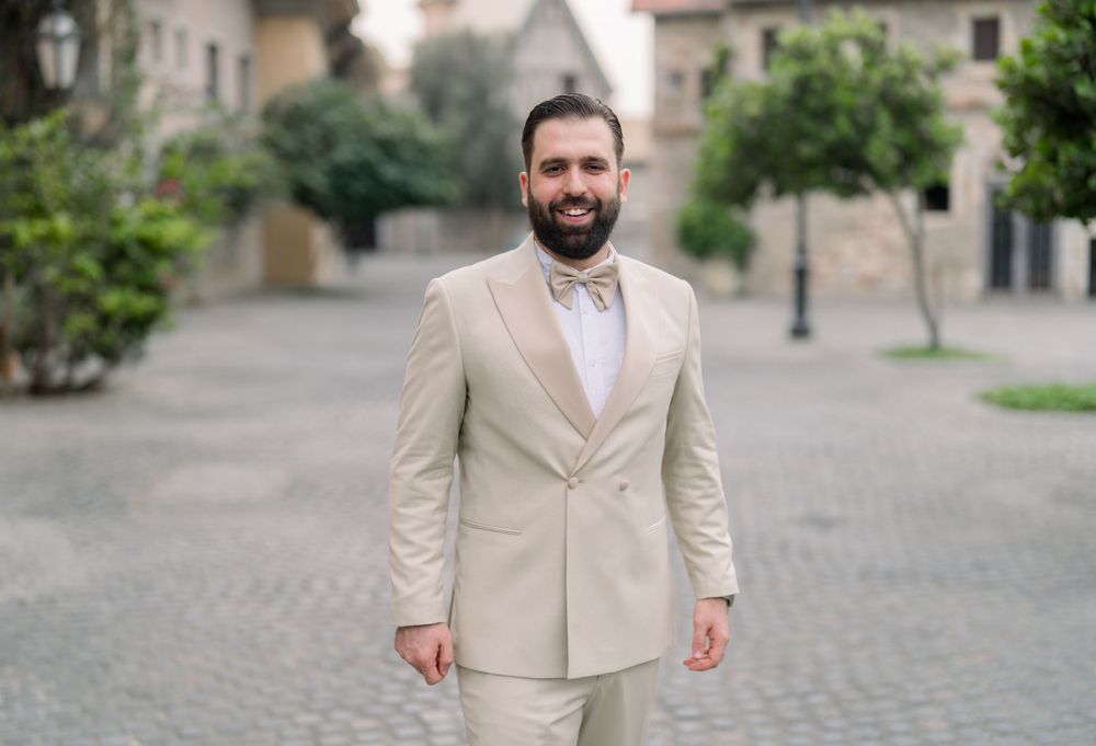 A person wearing a beige suit and bow tie stands smiling on a cobblestone street with buildings in the background.
