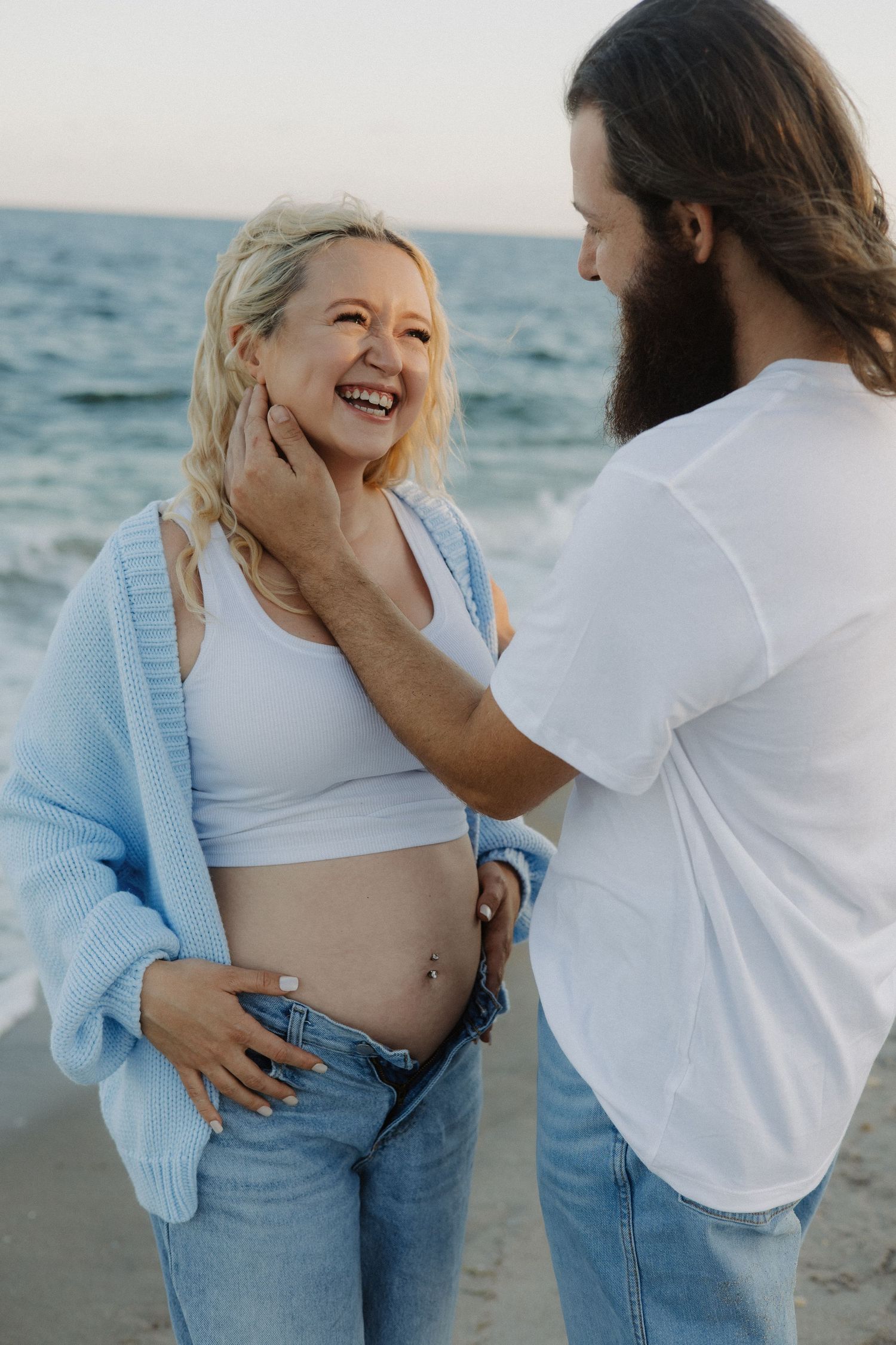 Pregnant women in ride lace dress holding her belly while looking down and smiling on the beach