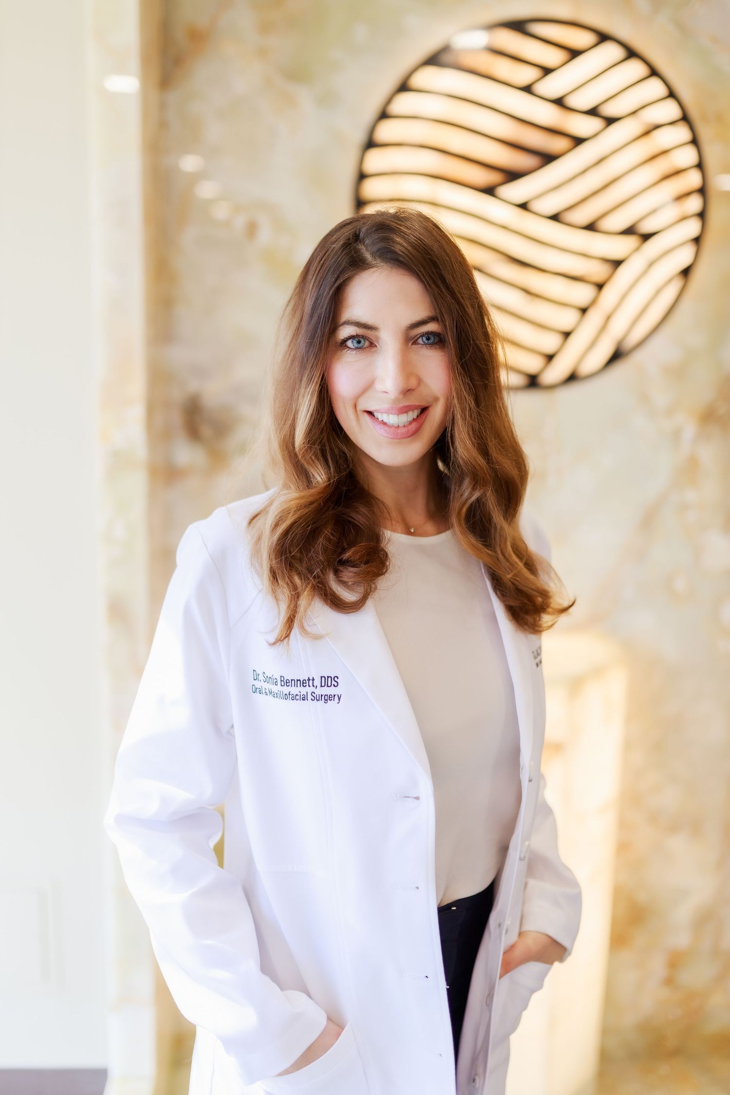 Healthcare professional in a white lab coat smiling in front of decorative wall lighting.