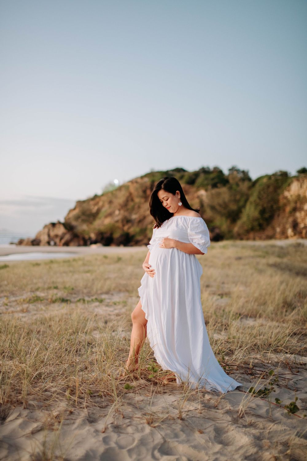 Expectant mother in flowing white dress poses gracefully on sandy beach with rocky cliffs in background.