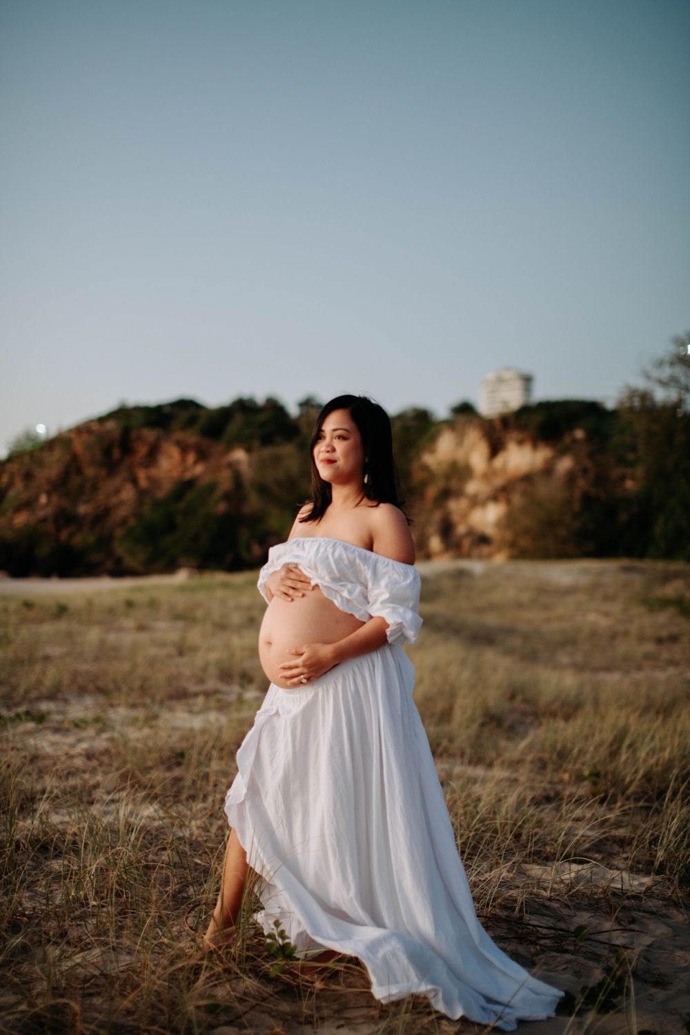 Pregnant woman in white off-shoulder gown stands against coastal landscape during golden hour.
