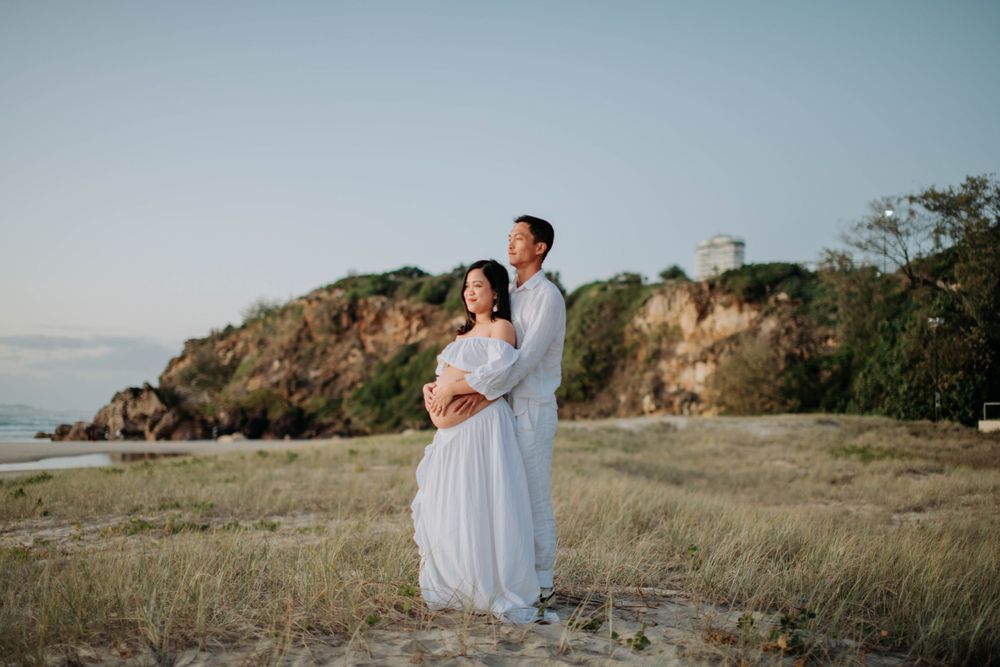 Couple in white attire share intimate moment during maternity photoshoot on coastal cliff.