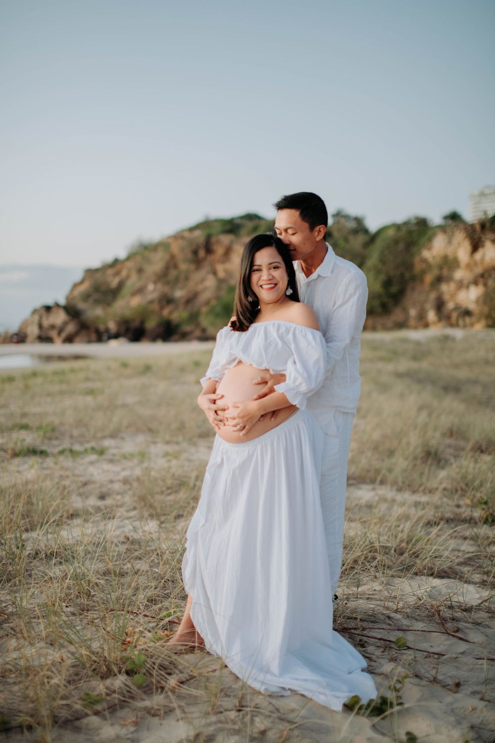 Romantic maternity portrait of couple in white clothing embracing on beach with cliffs behind.