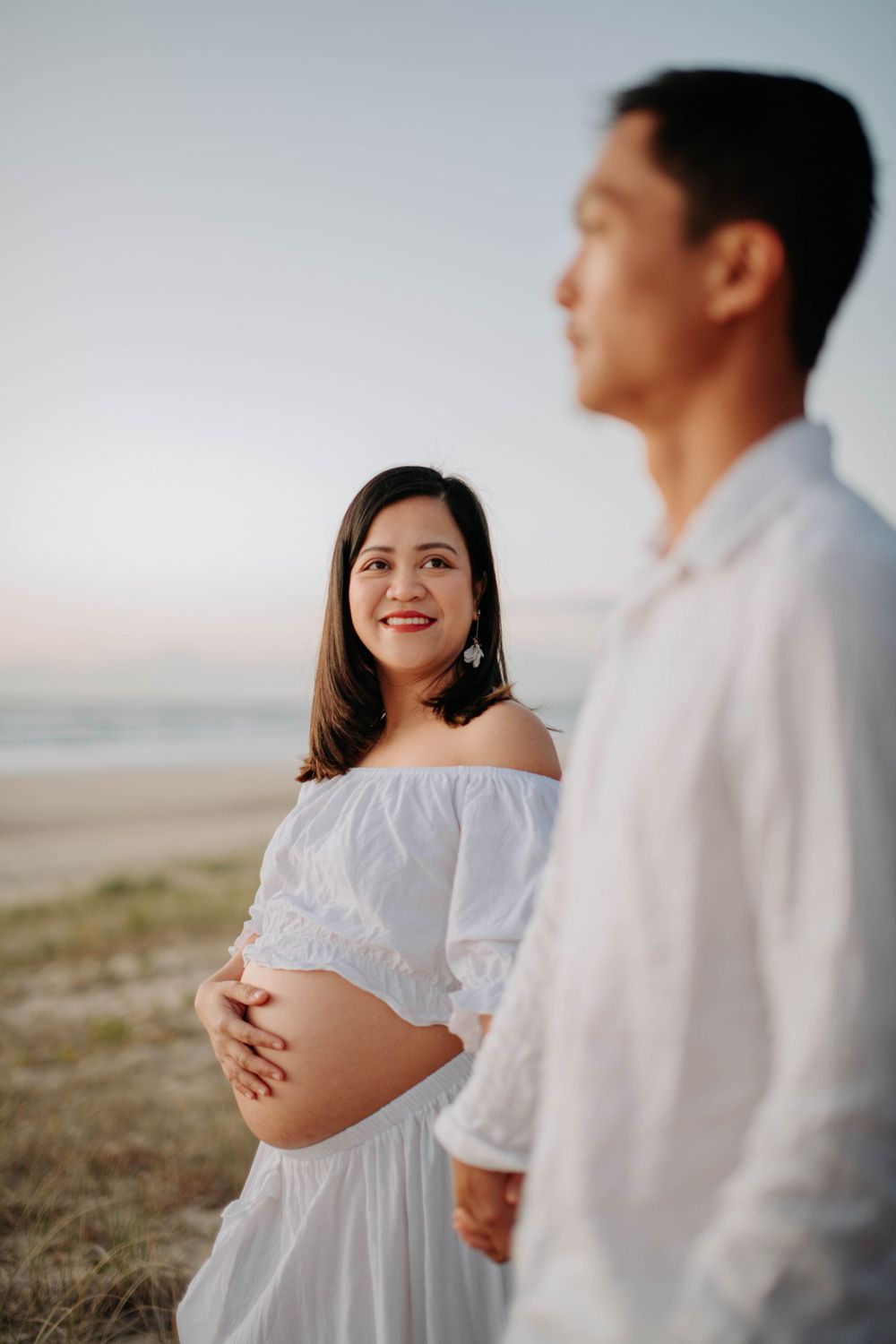 Loving couple in white outfits share tender moment during sunset maternity shoot at beach.