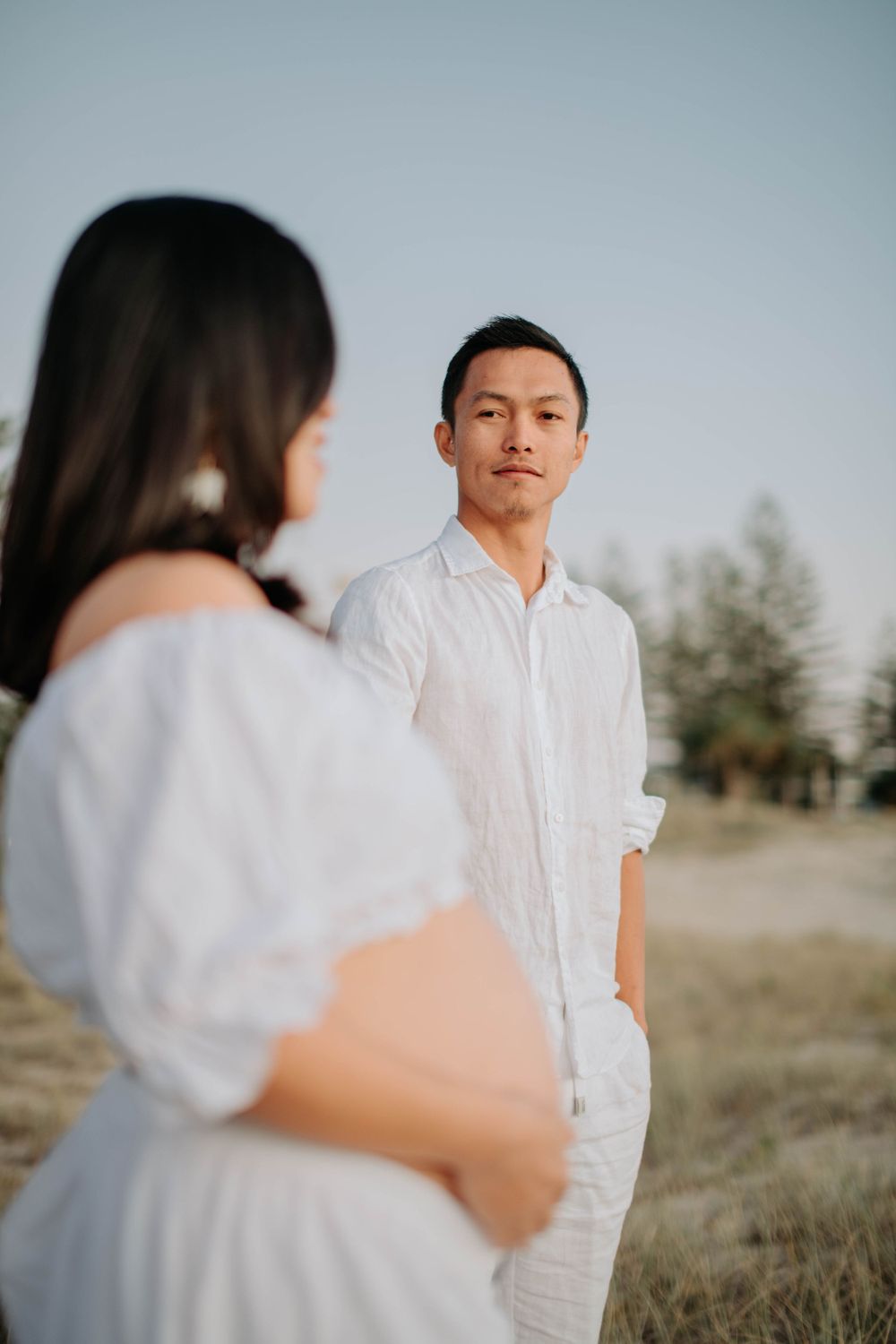 Intimate maternity portrait of couple in white clothing against natural outdoor setting.