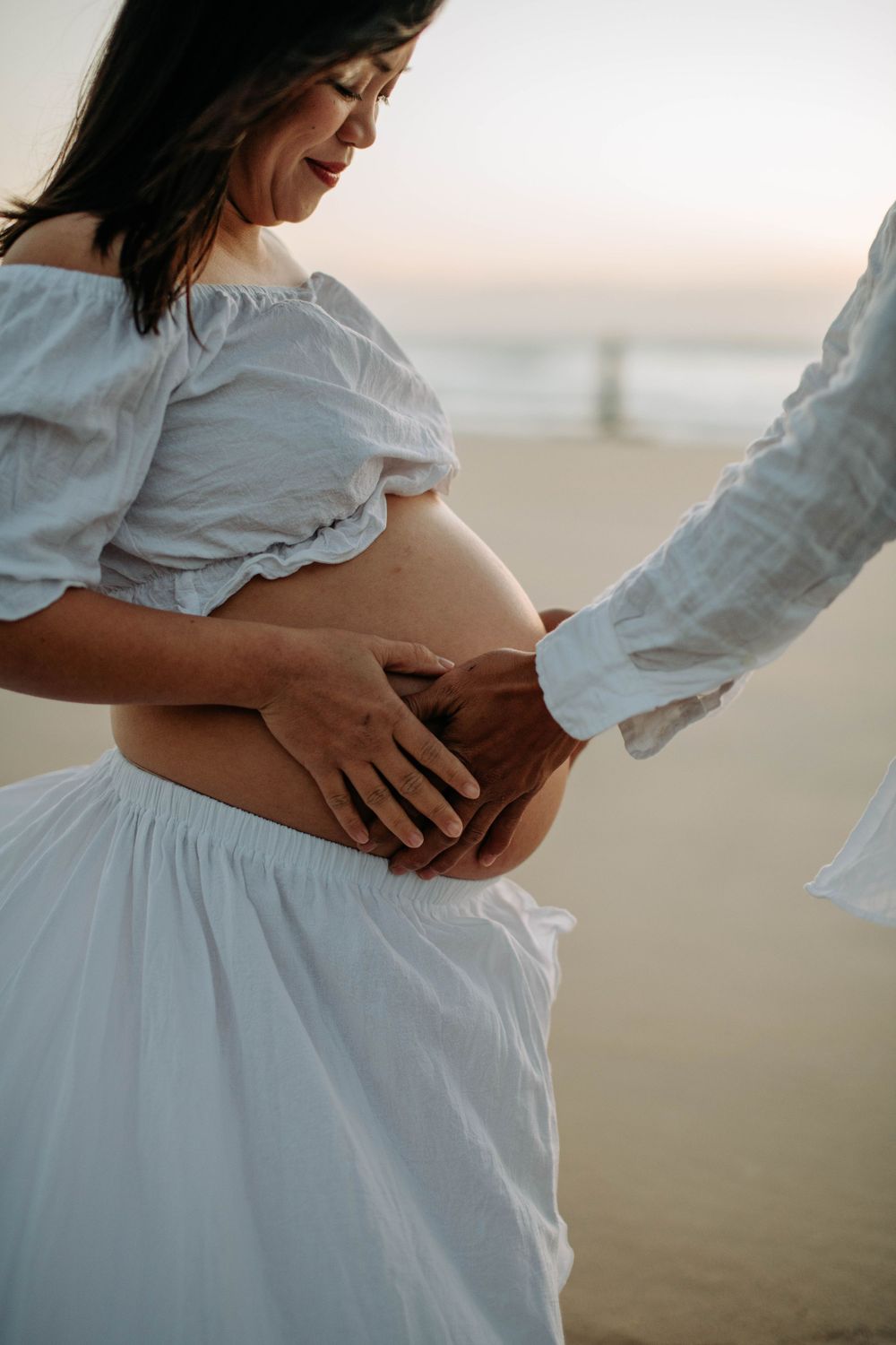 Intimate moment captured as hands cradle pregnant belly in white dress on beach.