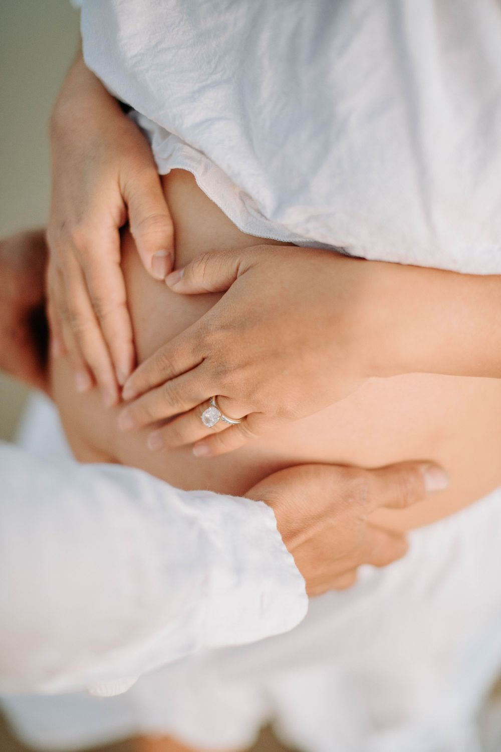 Close-up of hands forming heart shape on pregnant belly with engagement ring visible.
