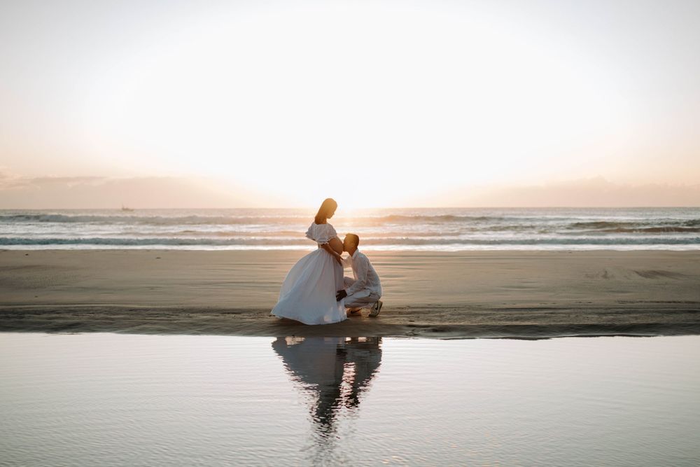 Couple creates silhouette against sunset beach reflection in romantic maternity pose.