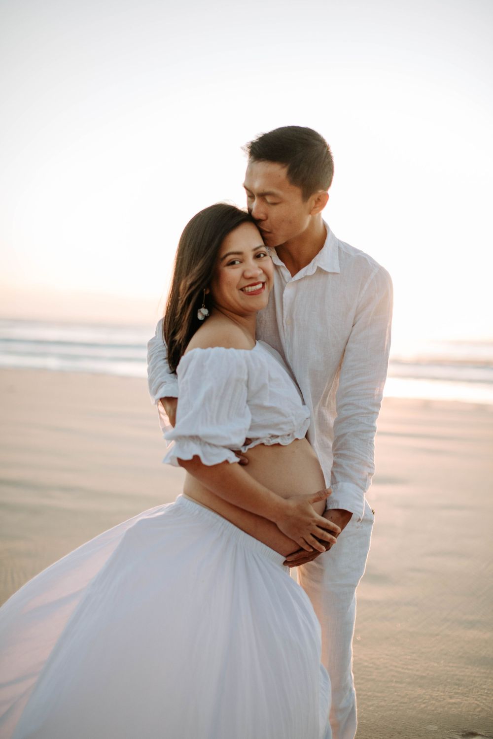 A romantic couple in white outfits embraces on the beach during sunset.