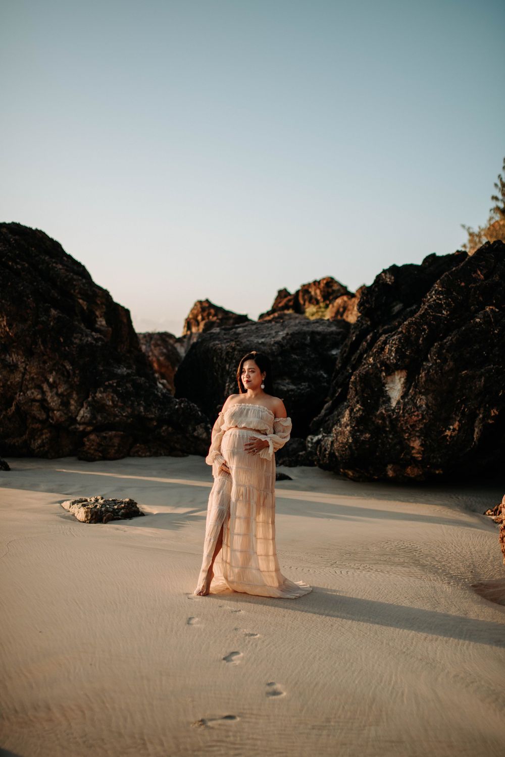 A maternity photo of an expectant mother in a flowy dress standing beside coastal rock formations.