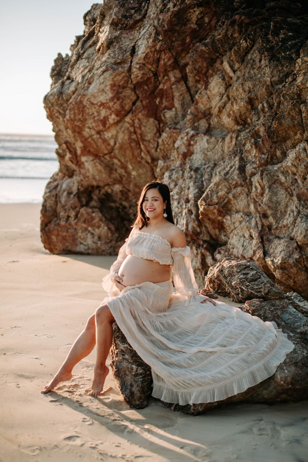 A beach maternity portrait showcasing a pregnant woman in white lace dress against rugged cliffs.