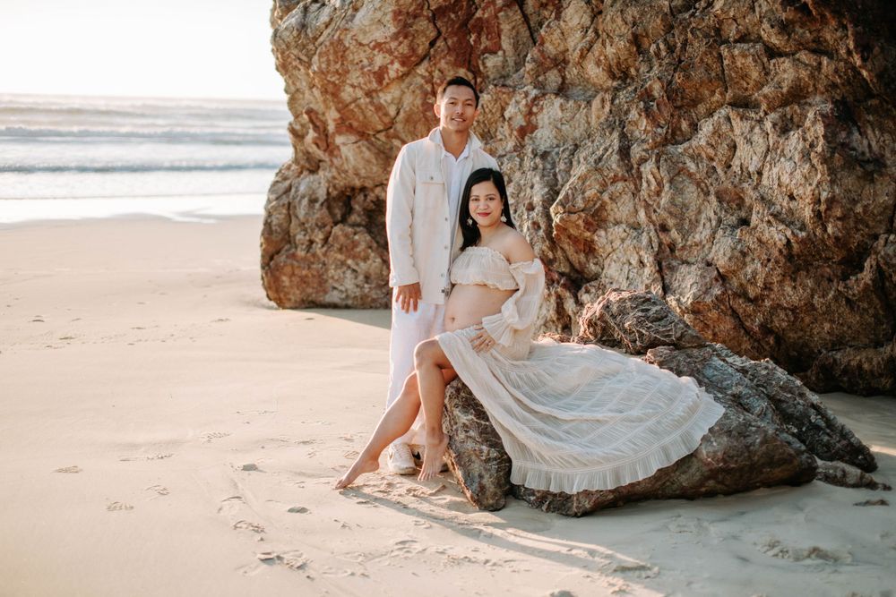 A beachside maternity photo featuring a couple posing near large coastal rocks.