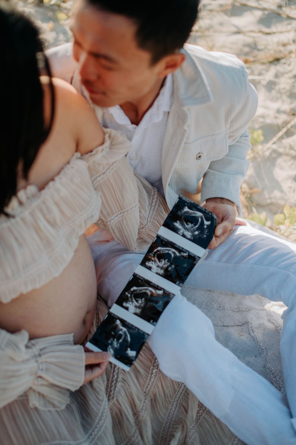 A couple shares an intimate moment while holding ultrasound photos on the beach.