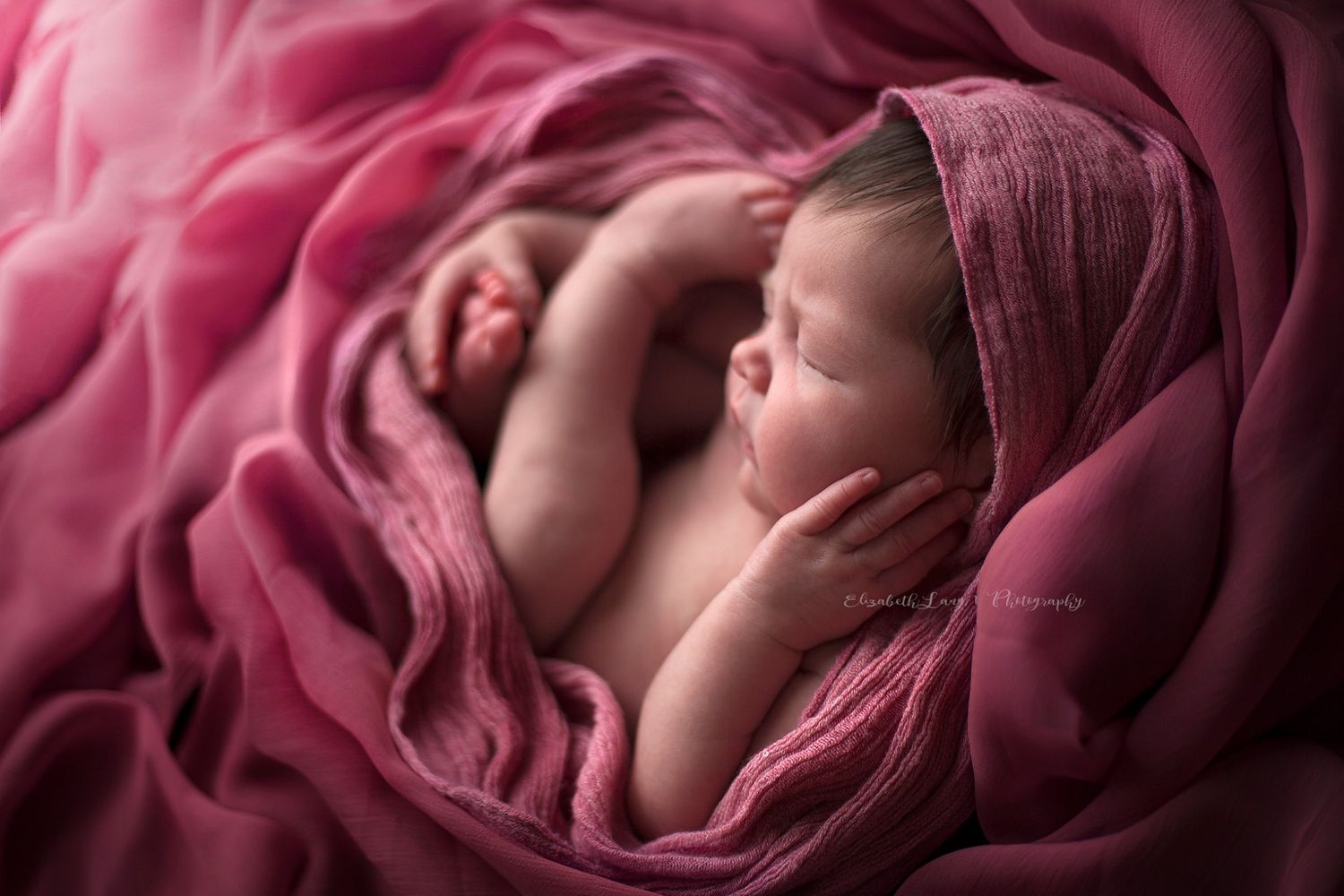 Newborn baby peacefully sleeping, swaddled in a soft, rose-colored wrap, surrounded by layers of flowing pink fabric. The baby is curled up