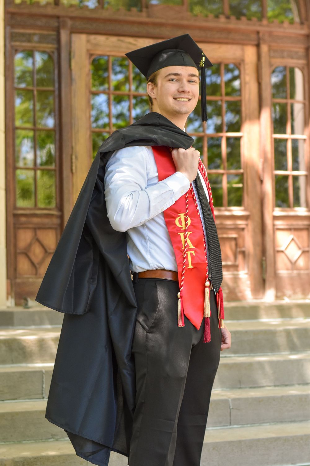 A graduate in cap and gown with a red and gold sash poses on campus steps in front of ornate wooden doors.