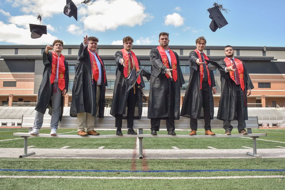 Six graduates in black robes and red stoles stand on stadium bleachers during graduation celebration outdoors.