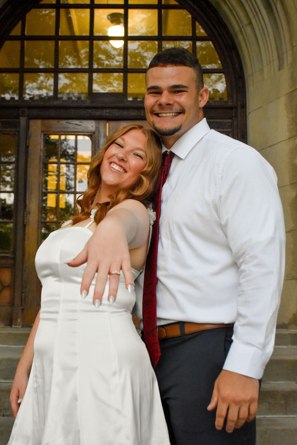 A couple poses in an archway entrance wearing white and gray attire while sharing a joyful moment together.