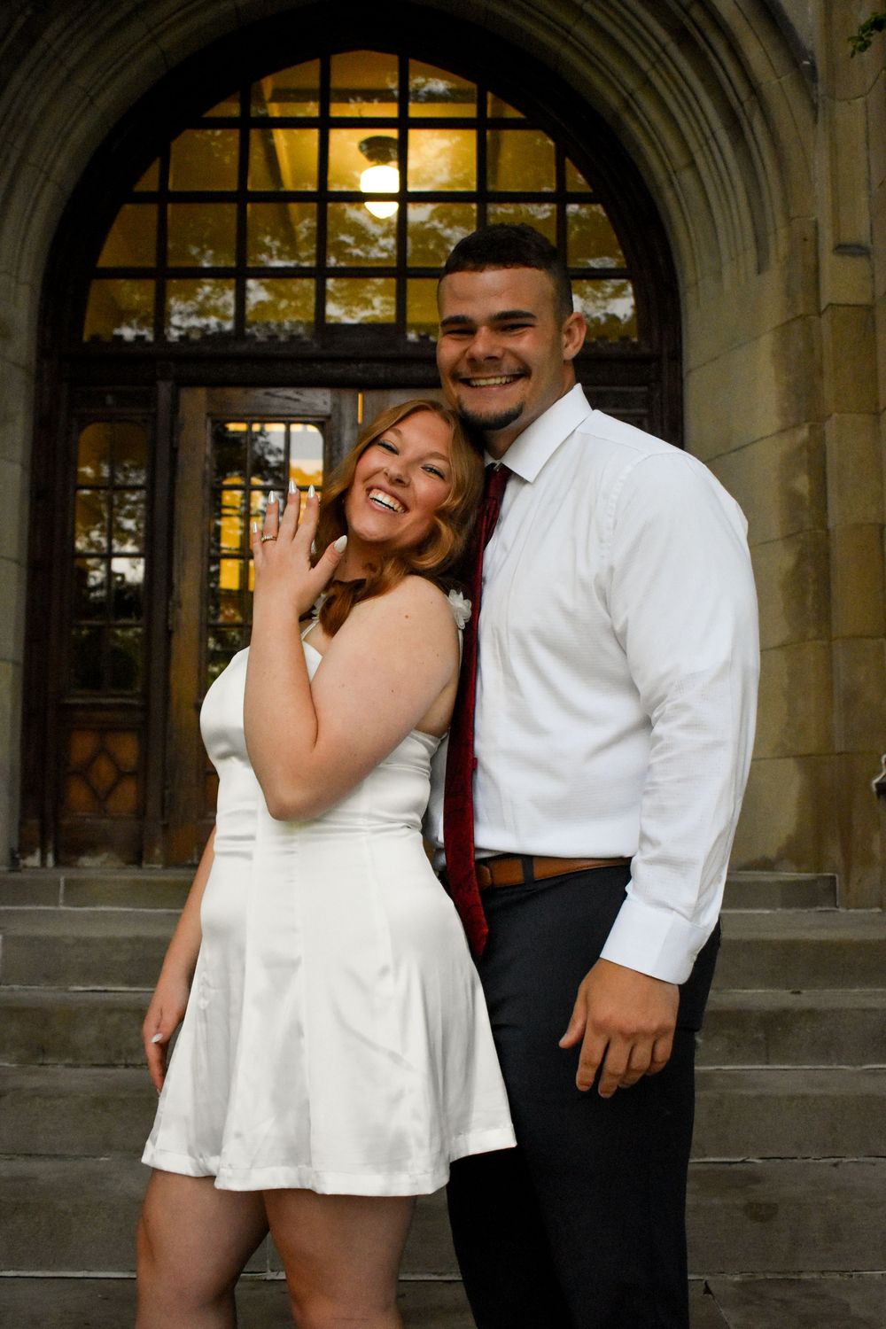 A couple in festive attire share a joyful moment together in front of an arched entrance with illuminated windows.