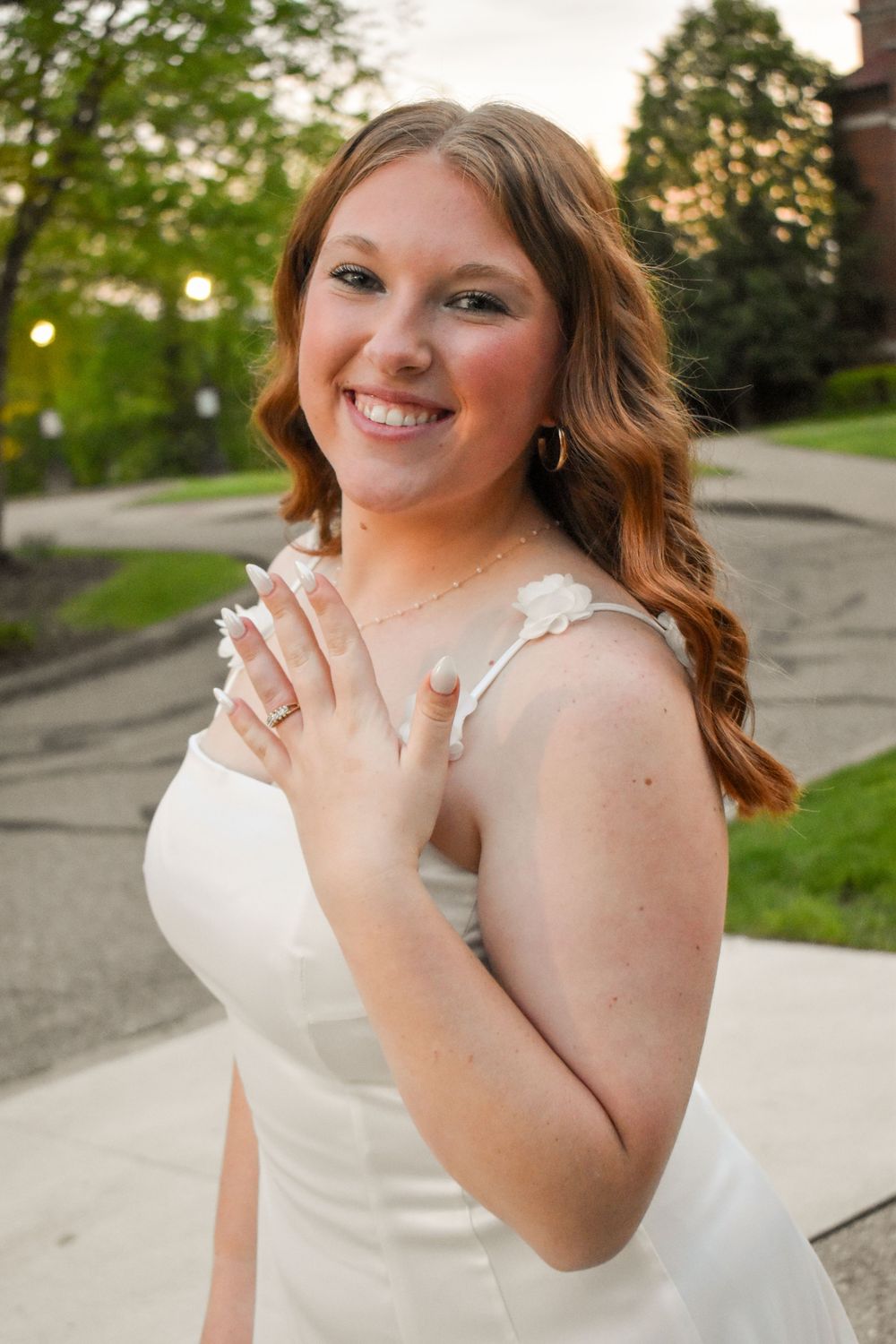 A person in a white sleeveless dress poses playfully with flowing hair against a natural outdoor backdrop.