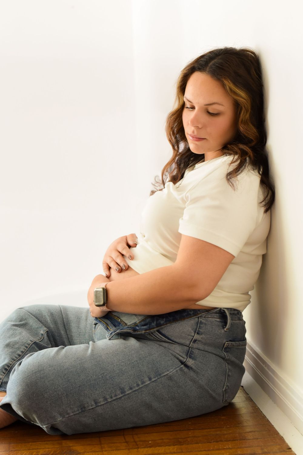 A person in a white t-shirt and jeans sits on a hardwood floor against a white wall in a contemplative pose.