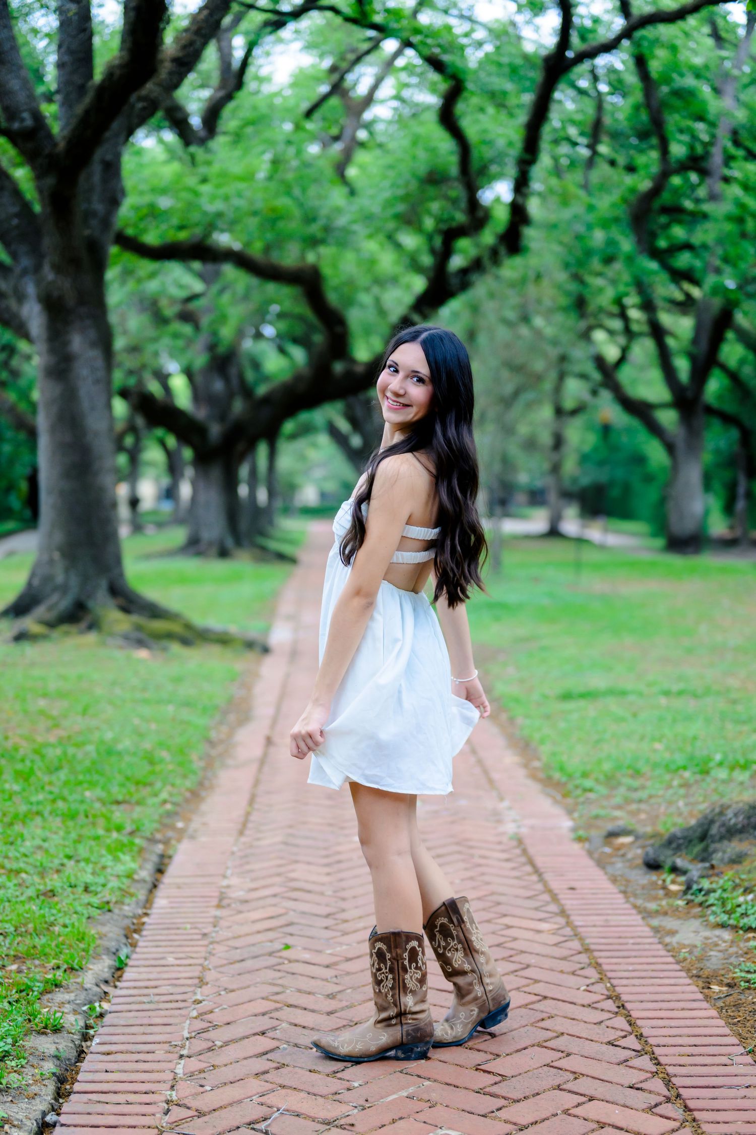 A figure in a white summer dress and brown boots poses playfully on a brick path surrounded by lush green trees in a park.