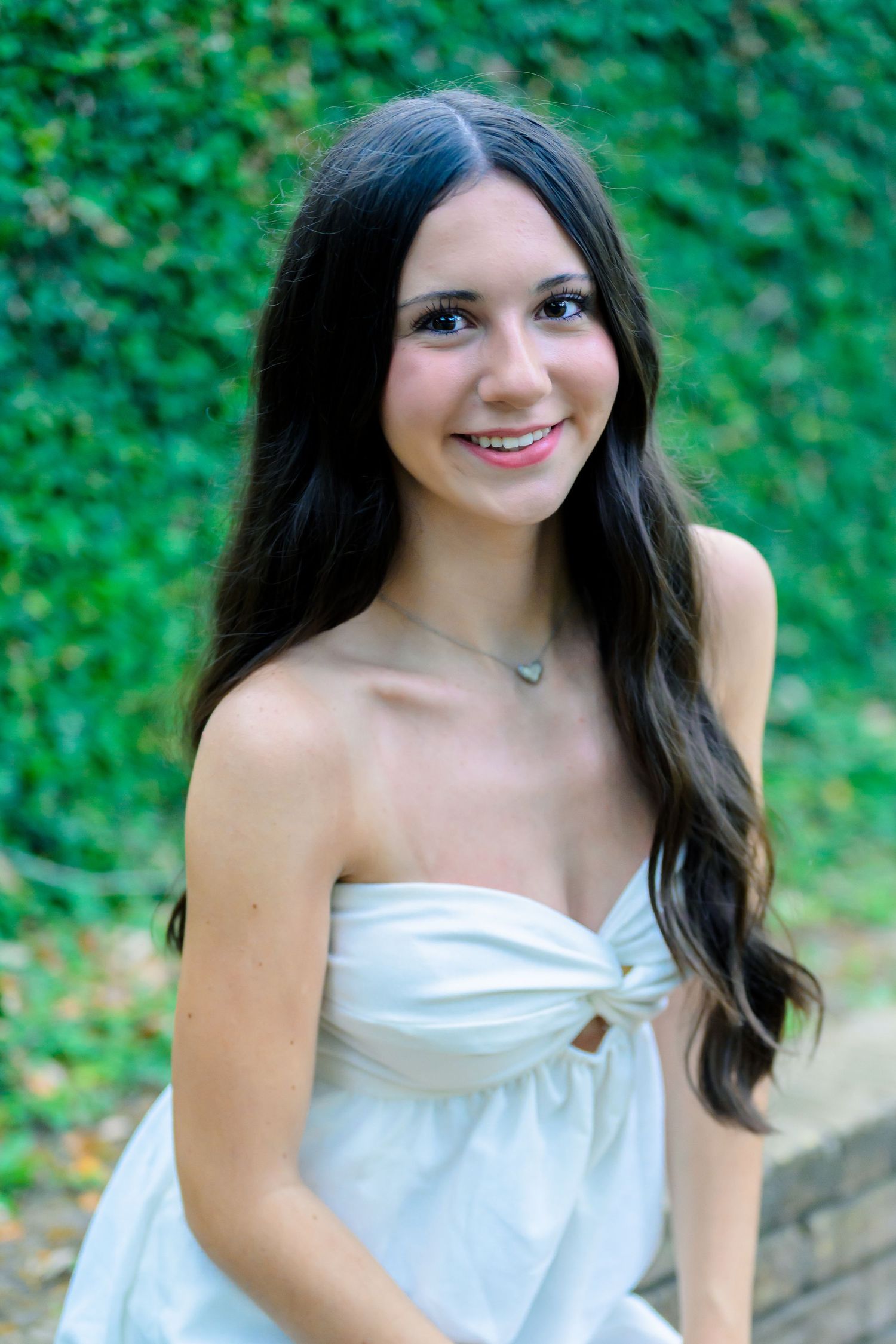 Portrait of someone wearing a white strapless dress with long dark hair and a necklace against a leafy green background.