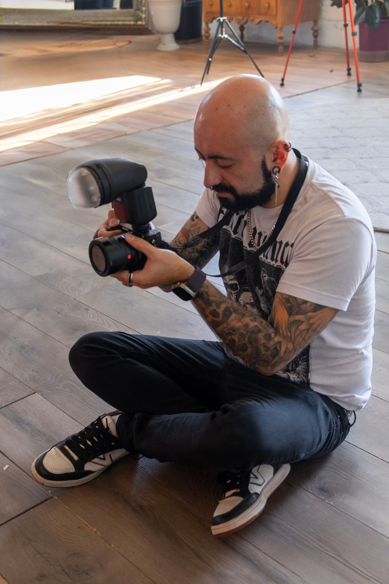 A tattooed photographer in a white t-shirt sits on a wooden floor while examining settings on a DSLR camera.