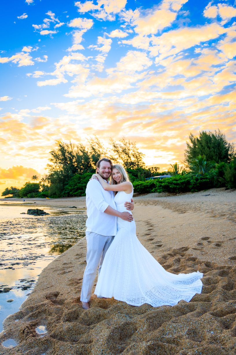 A romantic beach wedding photo at sunset with a couple embracing on sandy shore with dramatic sky and ocean view.