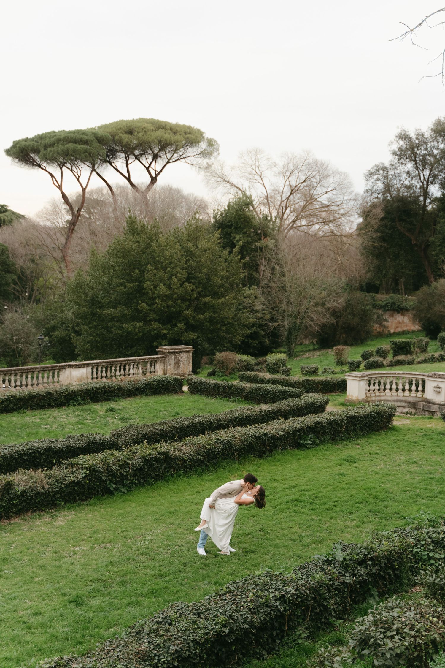 A bride in white bends back gracefully in a formal garden with manicured hedges and a stone balustrade.