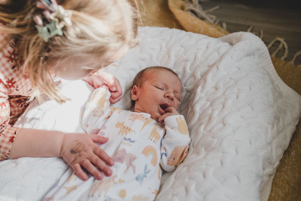 A sleeping infant lies on white bedding wearing a patterned sleeper in soft natural lighting.
