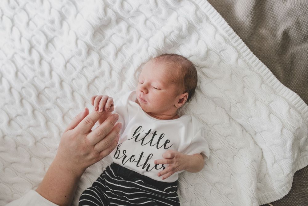 A newborn baby wearing a 'little brother' onesie lies on white bedding while holding an adult's hand.