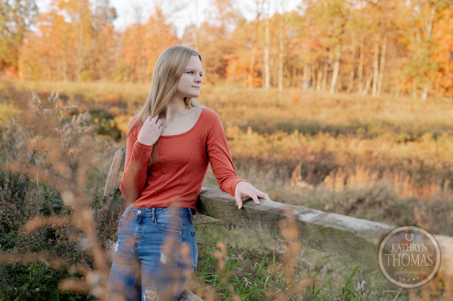 Person in orange top leans on rustic fence with fall foliage in background during sunset.