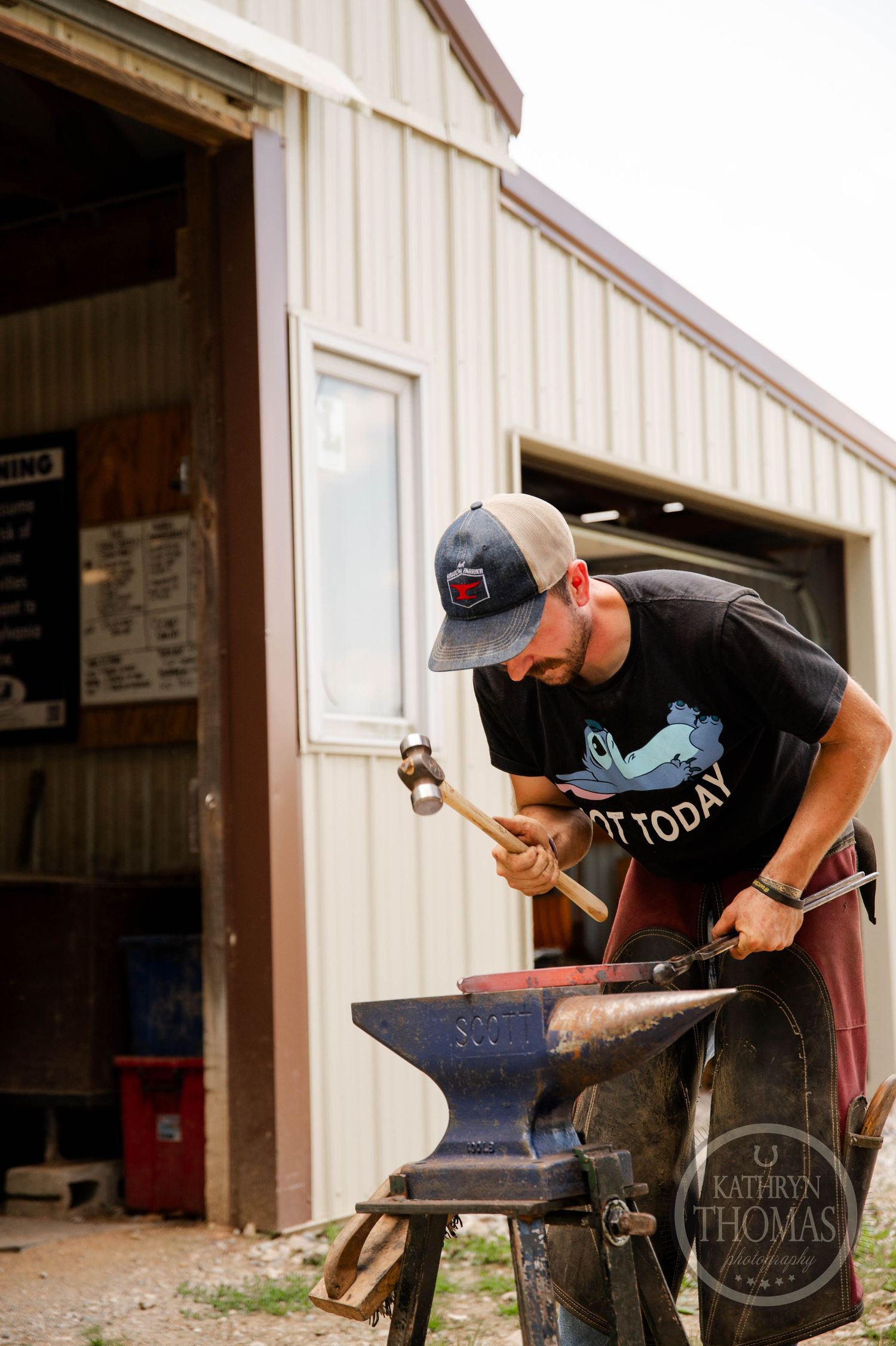 Farrier hammering at an outdoor anvil station beside the horse barn.