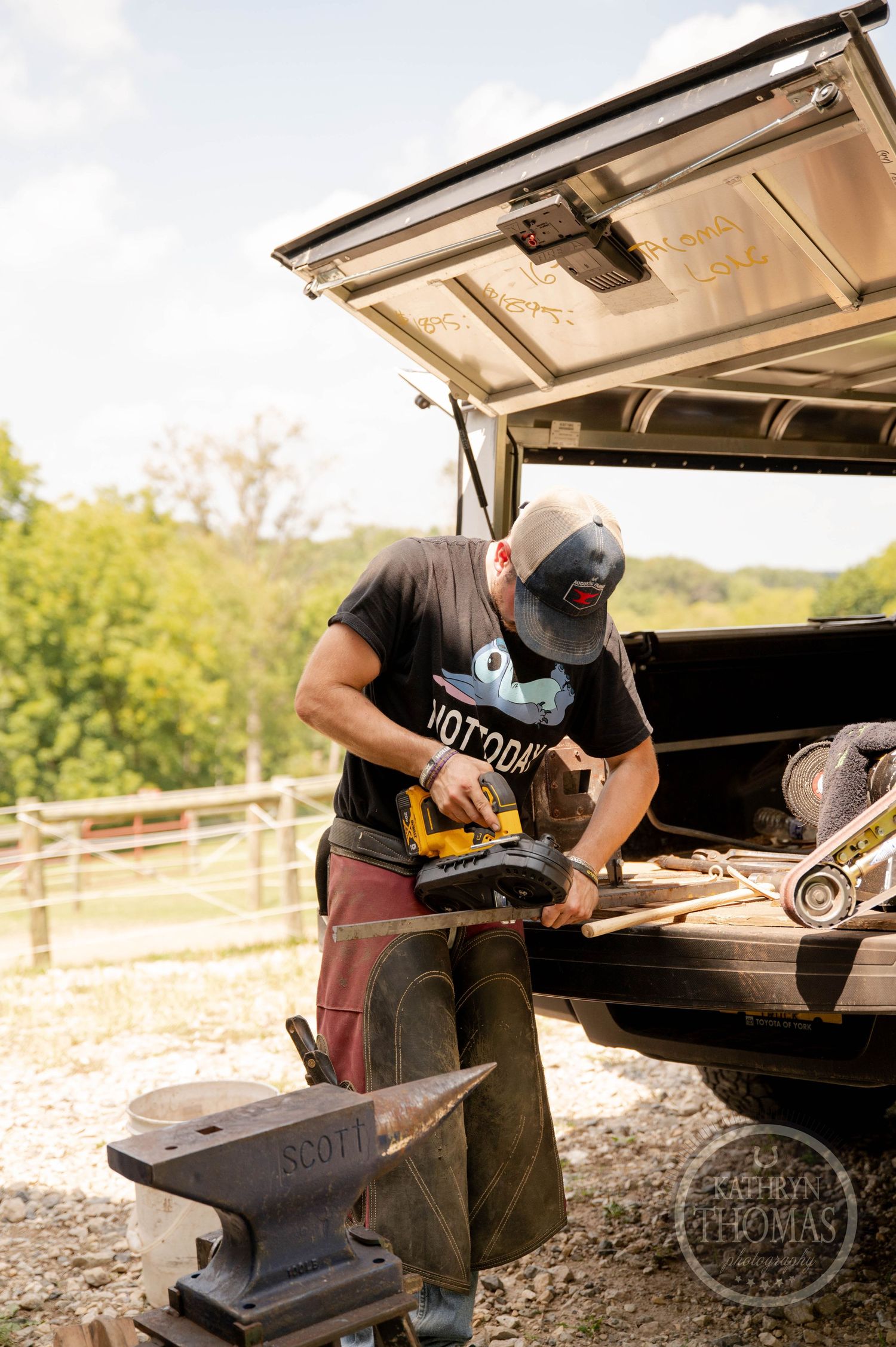 Farrier using tools at an anvil next to a truck with an open tailgate in outdoor setting.