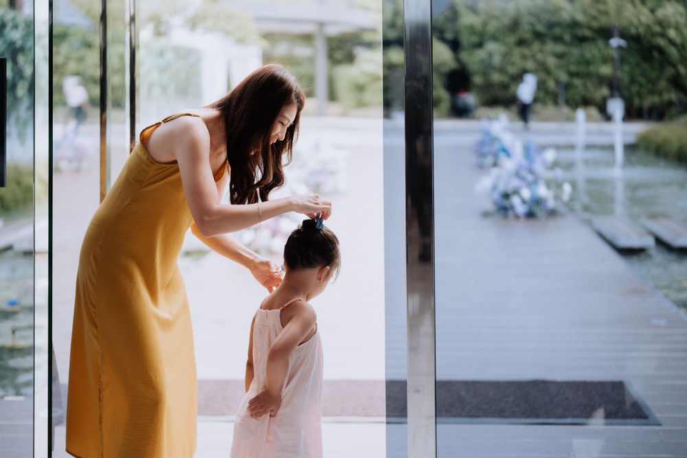Someone in a yellow dress leans down to fix a child's hair near a window with a blurred outdoor patio view.